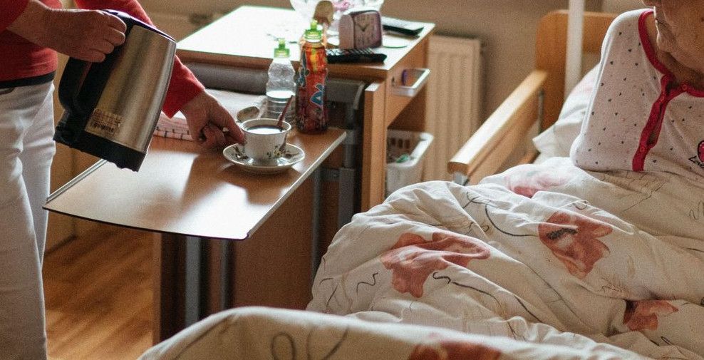 A caregiver serves tea to a senior woman in a cozy bedroom setting, fostering comfort and care.